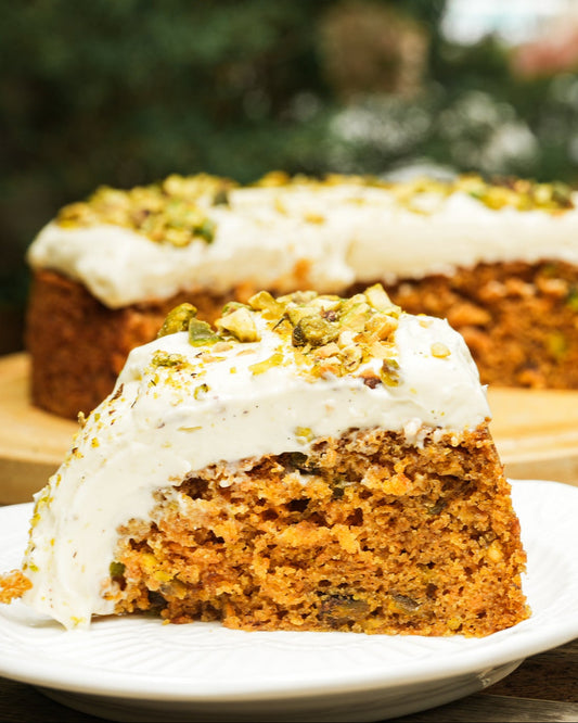 Carrot cake with white frosting on a plate, with the rest of the cake in the background.