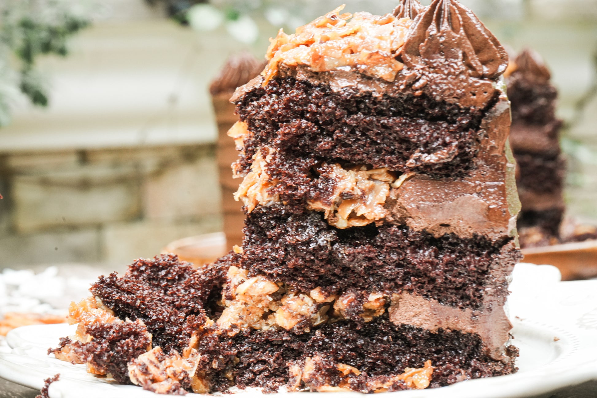 A close-up of a layered chocolate cake slice with rich dark brown layers, chocolate frosting, and caramel-like coconut pecan filling on a white plate.